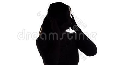 一个年轻快乐的女人戴着大耳机听音乐，拿着智能手机的剪影，女孩在白色的隔离巴上跳舞